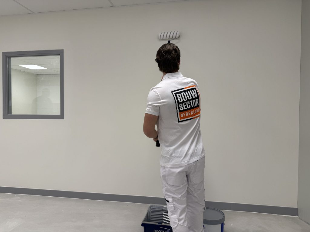 Schilder met roller en verfbak voor een lichte wand bij een binnenraam