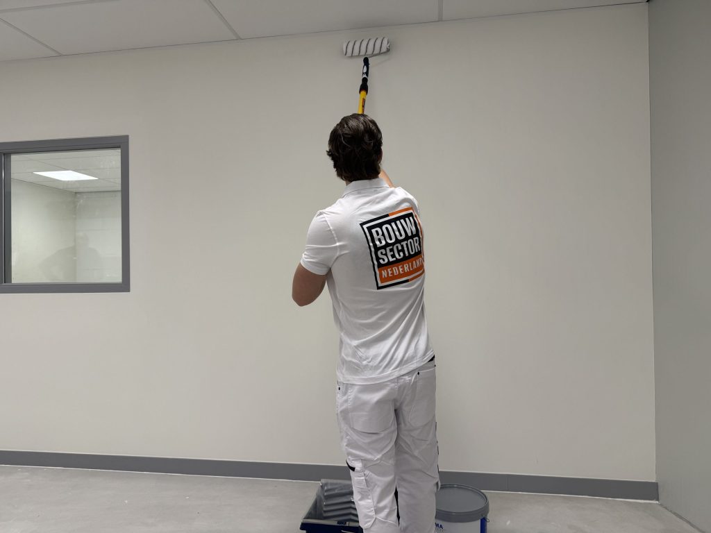 Schilder rolt met een lange steel de bovenrand van een lichte wand in een lege ruimte