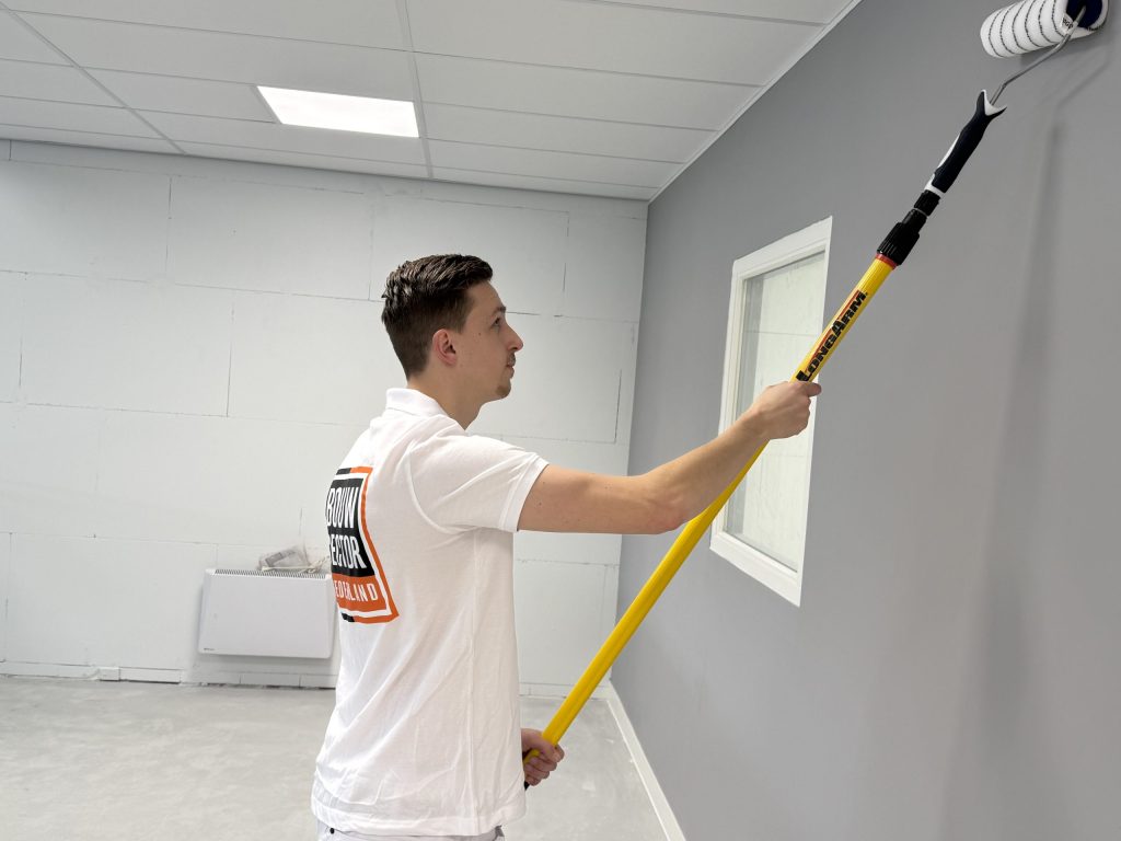 Schilder met verlengde roller verft een grijze muur naast een wit geschilderd raamkozijn