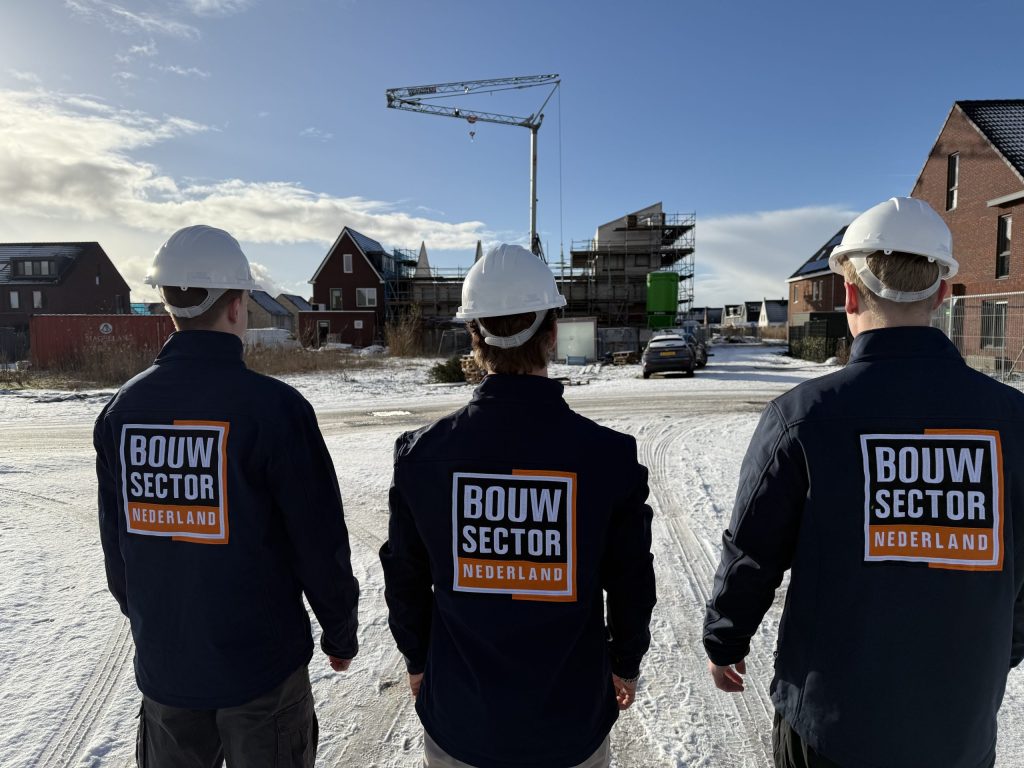 Drie vakmensen van Bouwsector Nederland in werkjas en witte helm lopen over een besneeuwde bouwplaats